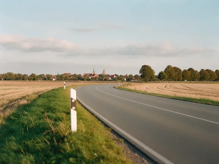 Landstraße zwischen abgeernteten Feldern, am Rand ein Leitpfosten, hinten ein Dorf mit Kirchturm